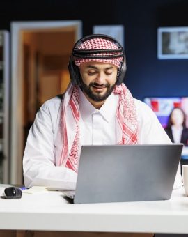 Professional Arab guy at his desk, diligently using his laptop for research. He engages in video conferencing and email communication, showcasing a blend of traditional attire and wireless technology.