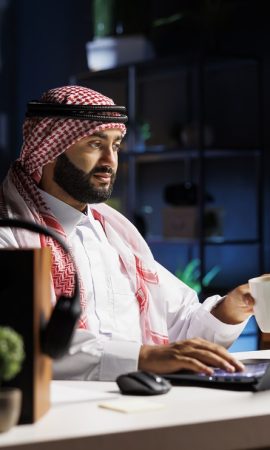 Side-view of a Muslim guy holding a cup of coffee and surfing the net on his digital laptop. Image showcasing a male Arab person engrossed in his personal computer while drinking tea.