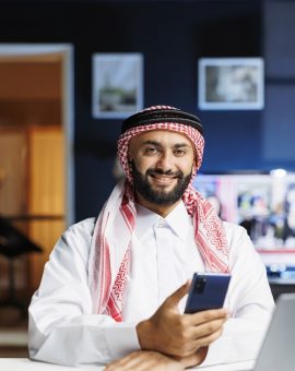 Male Arab person in profile, grinning at camera with his personal computer close by. Using contemporary technology, smiling Muslim guy with a cell phone, portrayed in portrait with laptop on table.